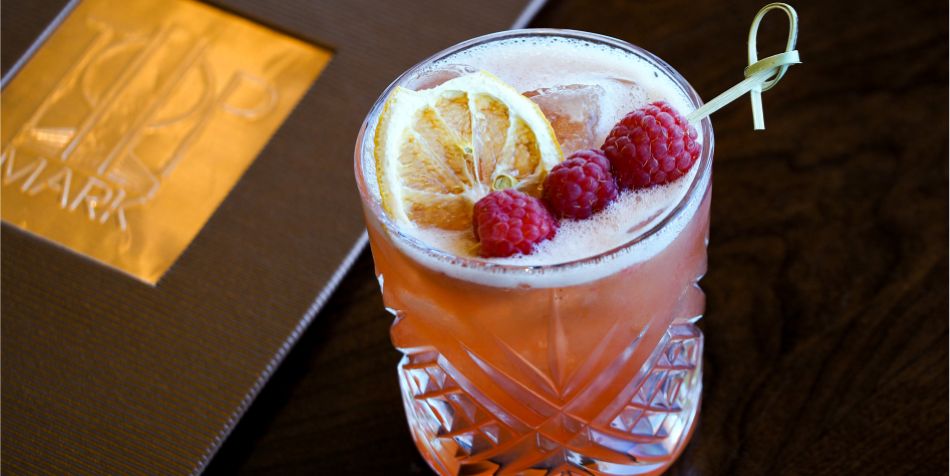 Pink cocktail garnished with dried citrus and fresh raspberries, served in a patterned glass beside a Top of the Mark menu.