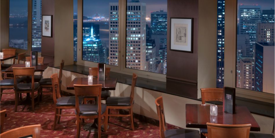 Evening dining area at Top of the Mark with wooden tables set against large windows showcasing San Francisco city lights at night.