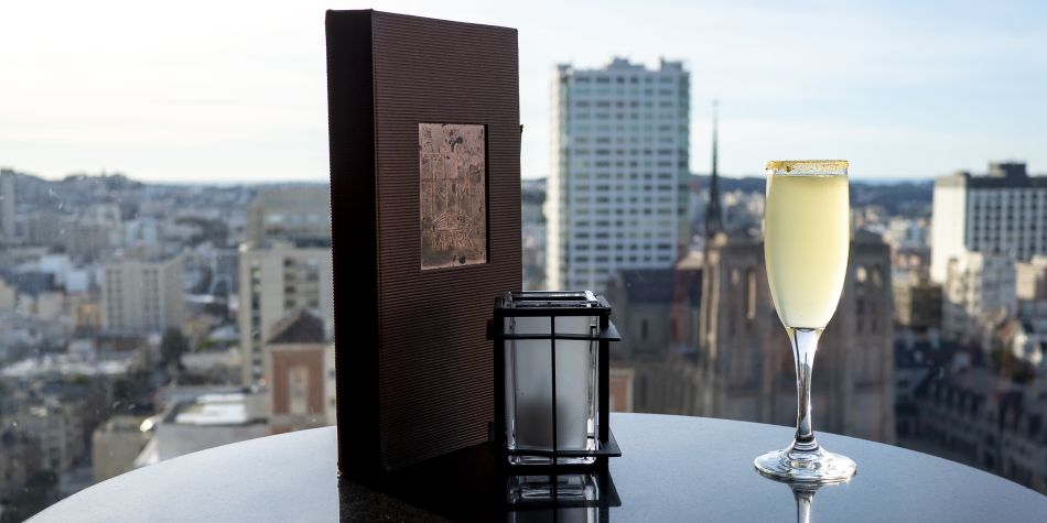 Sparkling cocktail in a flute beside a Top of the Mark menu and candle on a table, with city skyline views through the window.