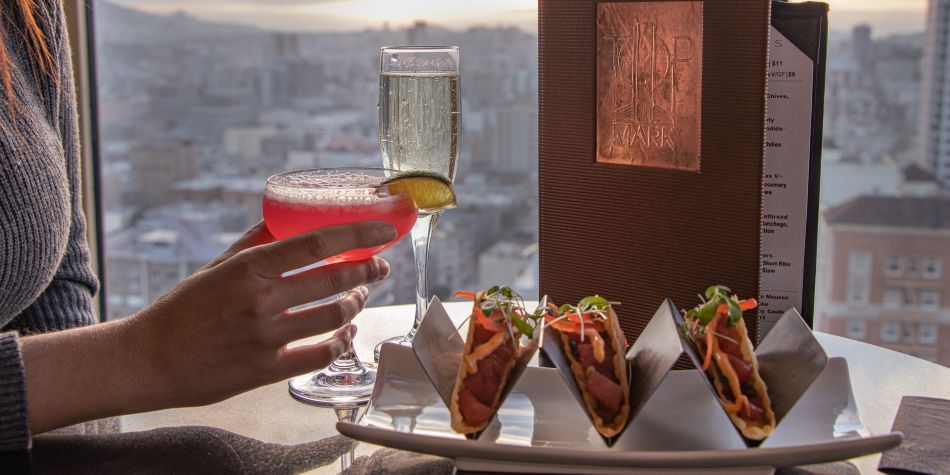 Cocktails and tacos served by a window, with city views in the background and a Top of the Mark menu standing beside the table.