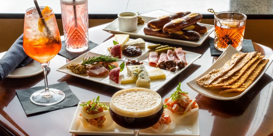 Assortment of cocktails and shared plates, including charcuterie, crackers, appetizers, and desserts, arranged on a table by a window with city views.