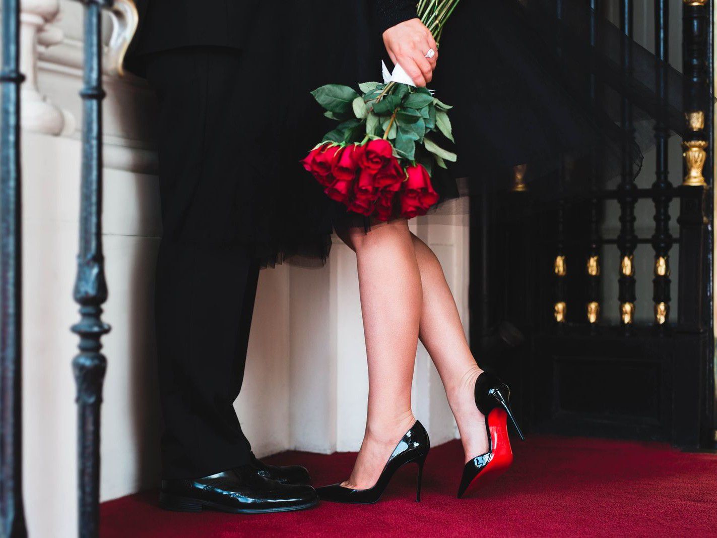 Two people standing close together, one holding a bouquet of red roses on a red carpeted entrance.