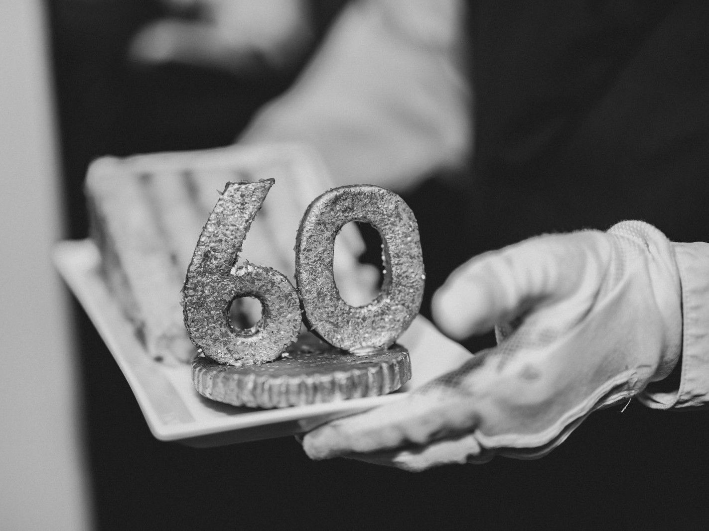 A tray holding a candle shaped as the number 60, presented by a gloved hand.
