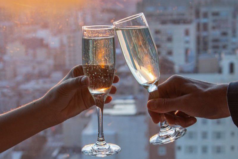 Two hands clinking champagne glasses in front of a city skyline at sunset.