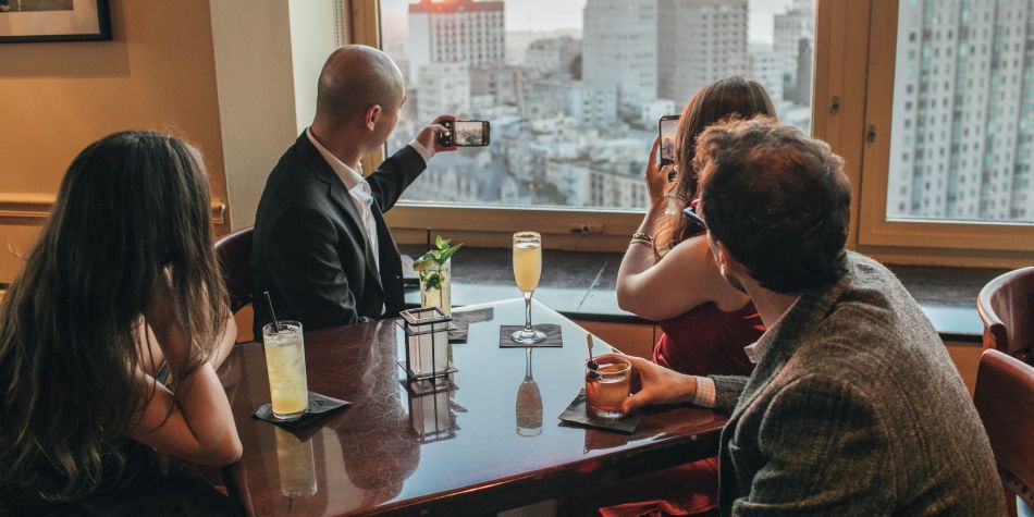 Group of guests seated at a table by a window, enjoying cocktails while taking photos of the city skyline view.