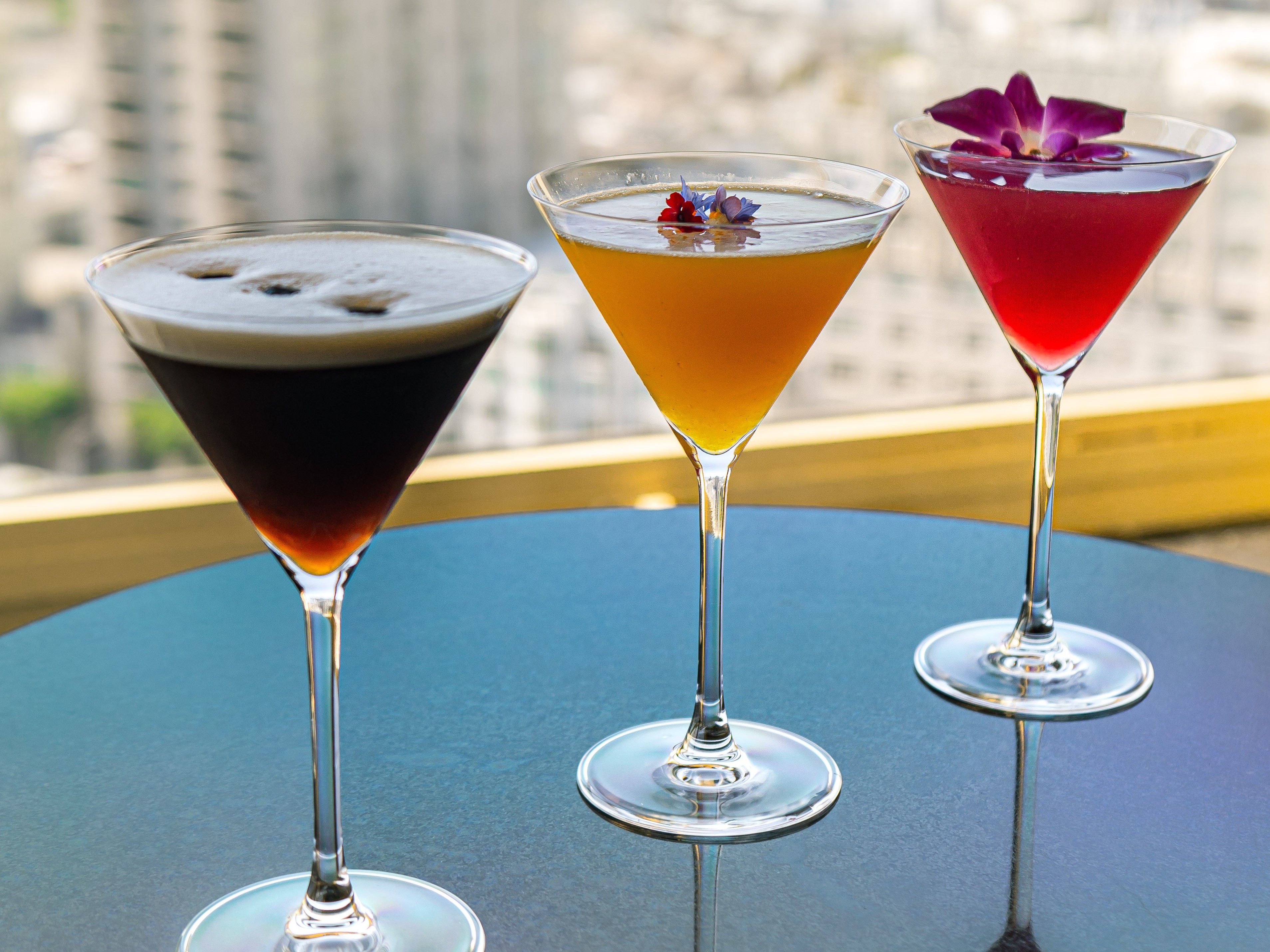 Three martini glasses on a table, including a dark espresso-style cocktail, an amber-colored drink with small flower garnish, and a red cocktail with a purple flower, with a blurred city view in the background.
