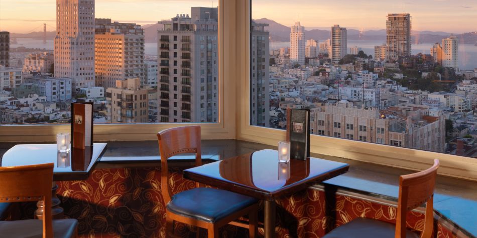 Window-side tables at Top of the Mark overlooking the city skyline and bay at sunset, with menus and candlelit tables set for guests.