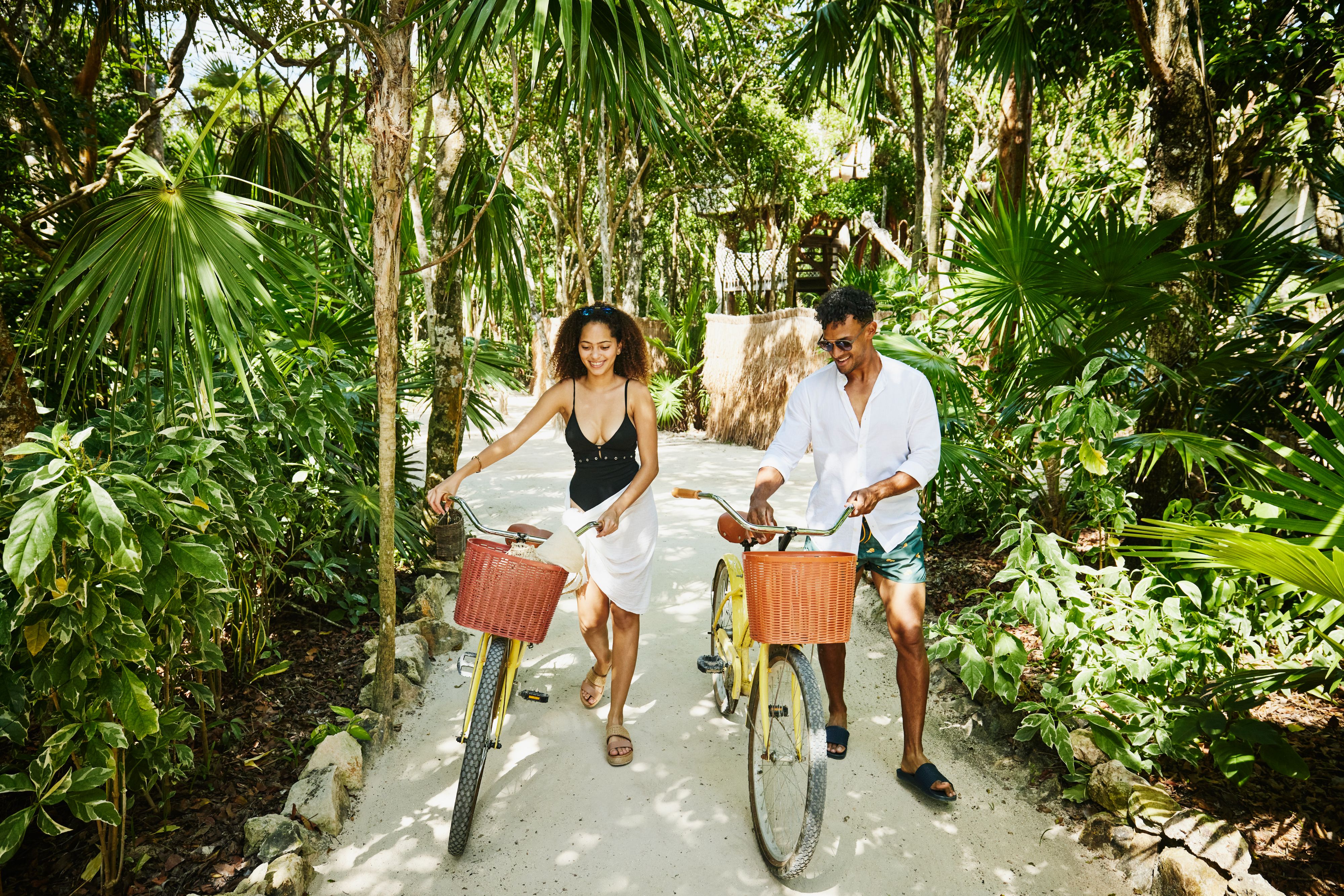 Guests walk bicycles along a shaded tropical pathway lined with lush palm trees and greenery, exploring the resort’s natural surroundings on a sunny day.