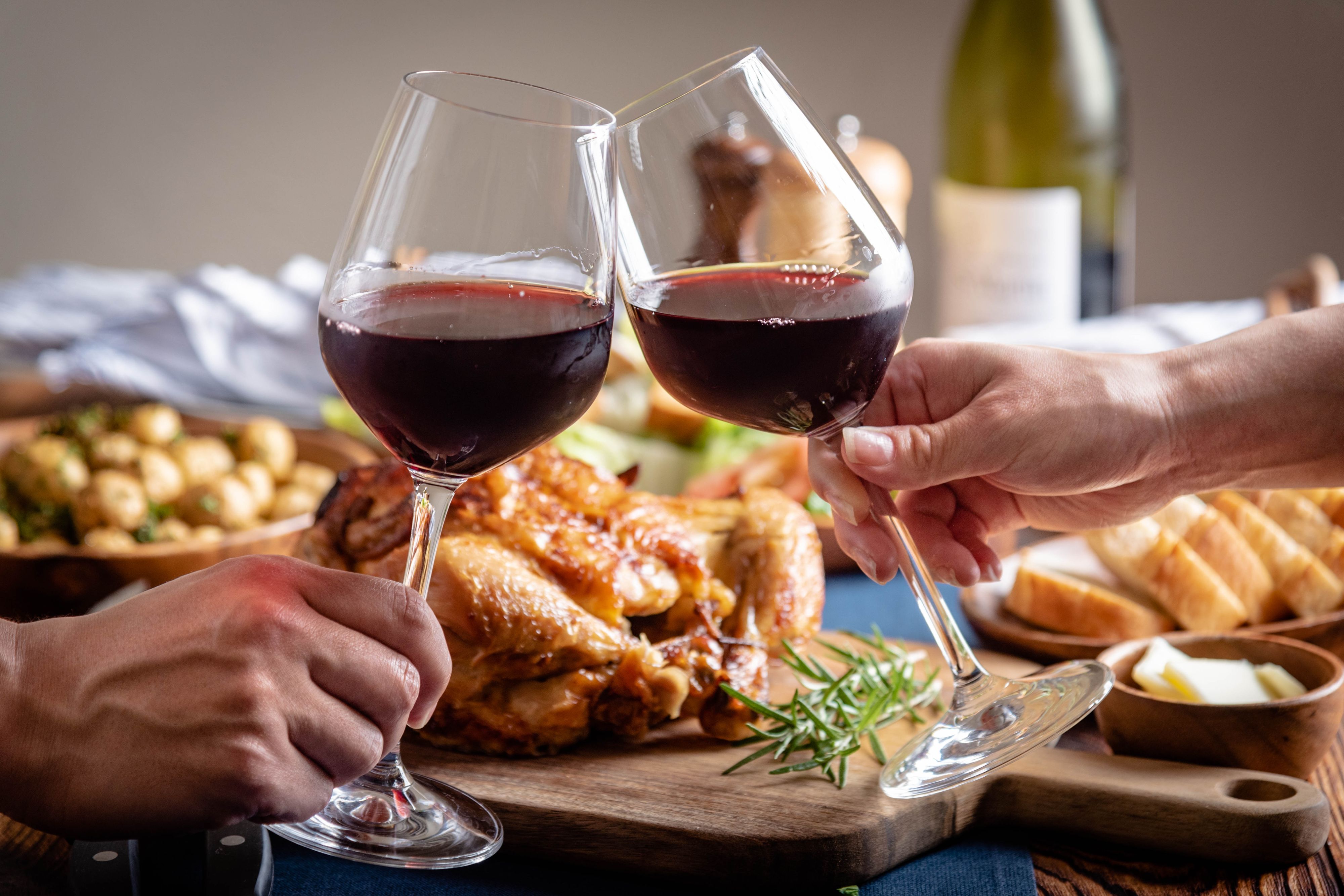 Two glasses of red wine meet in a toast over a rustic Thanksgiving table, with a golden roast turkey, rosemary, and warm autumn dishes.