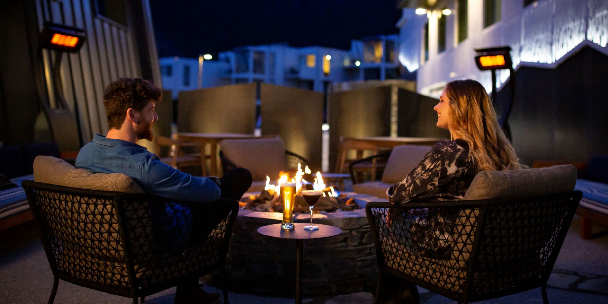 Man and woman sitting in front of a bonfire and smiling at each other