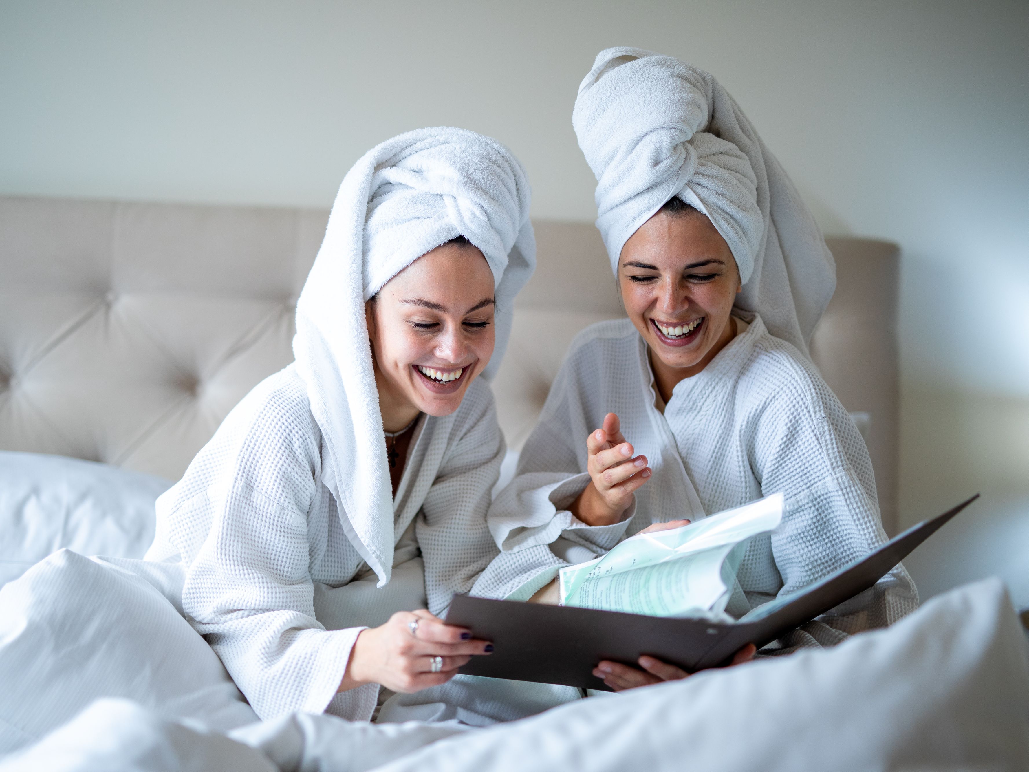 Two women in white robes sitting on hotel bed looking at a menu.