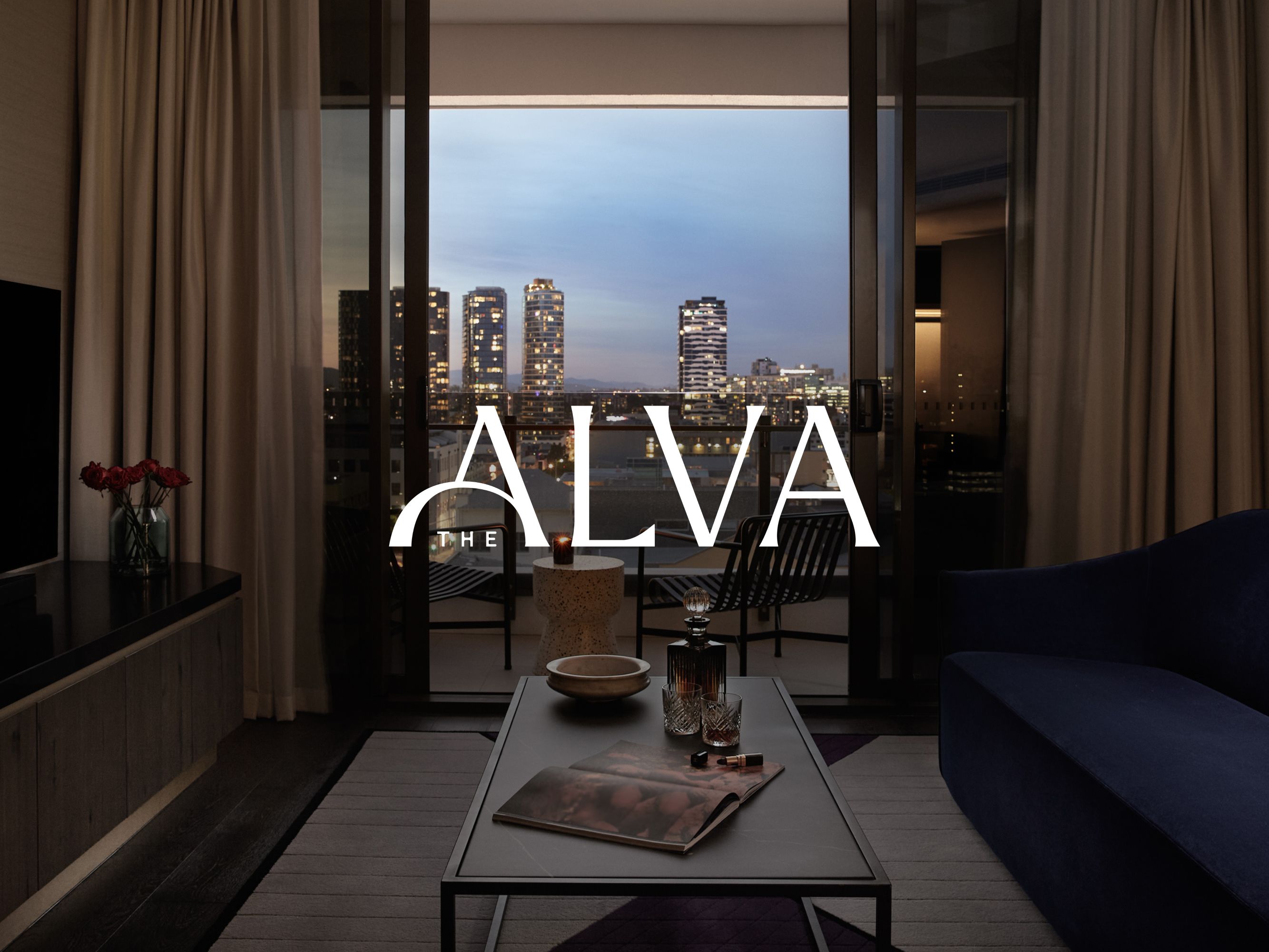 Logo properti berwarna putih ditampilkan di atas foto interior kamar yang bernuansa temaram di The Alva Hotel Brisbane dengan pemandangan kota di latar belakang.