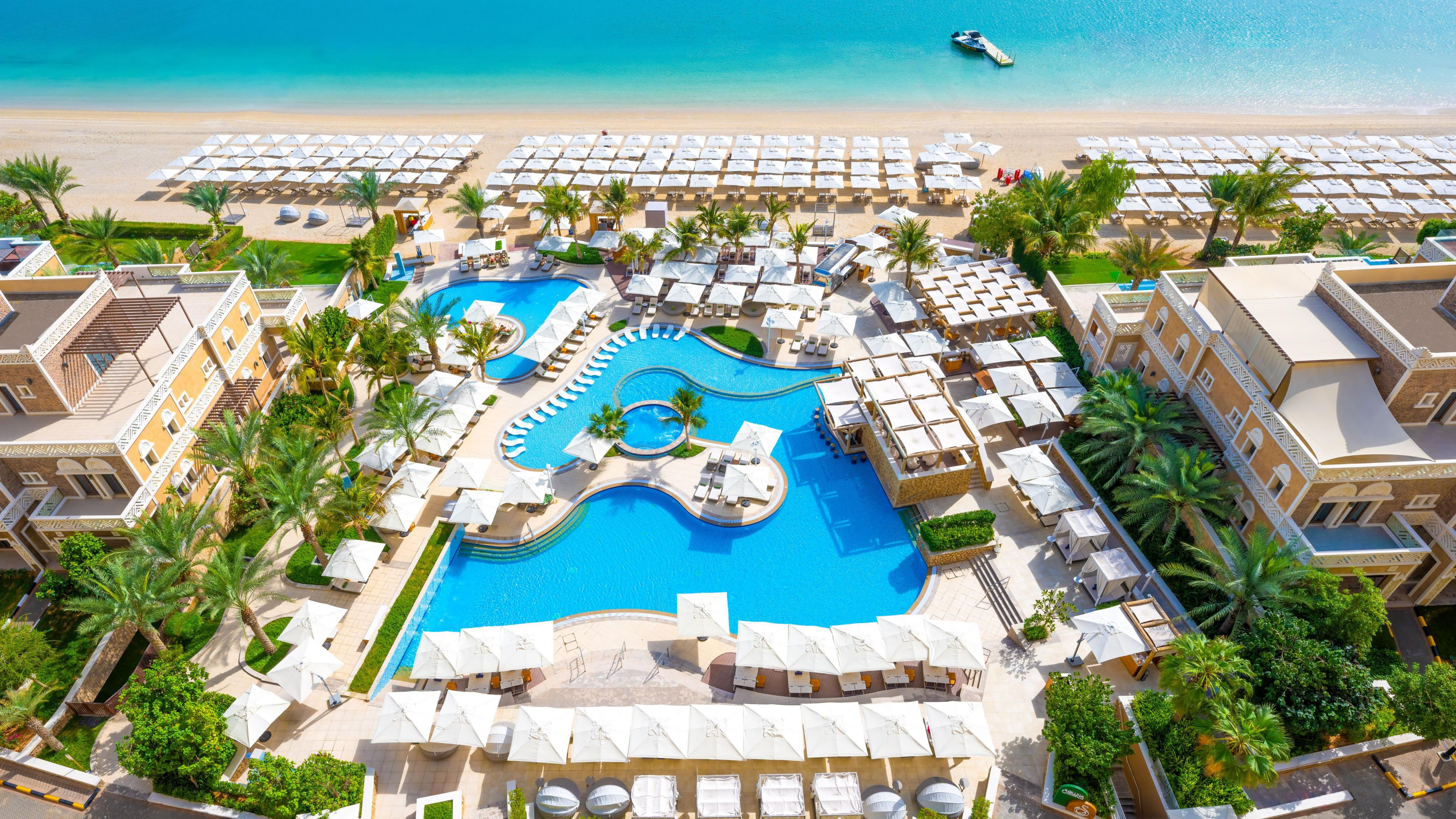 A large, central blue swimming pool with multiple sections, surrounded by numerous sun loungers and white umbrellas