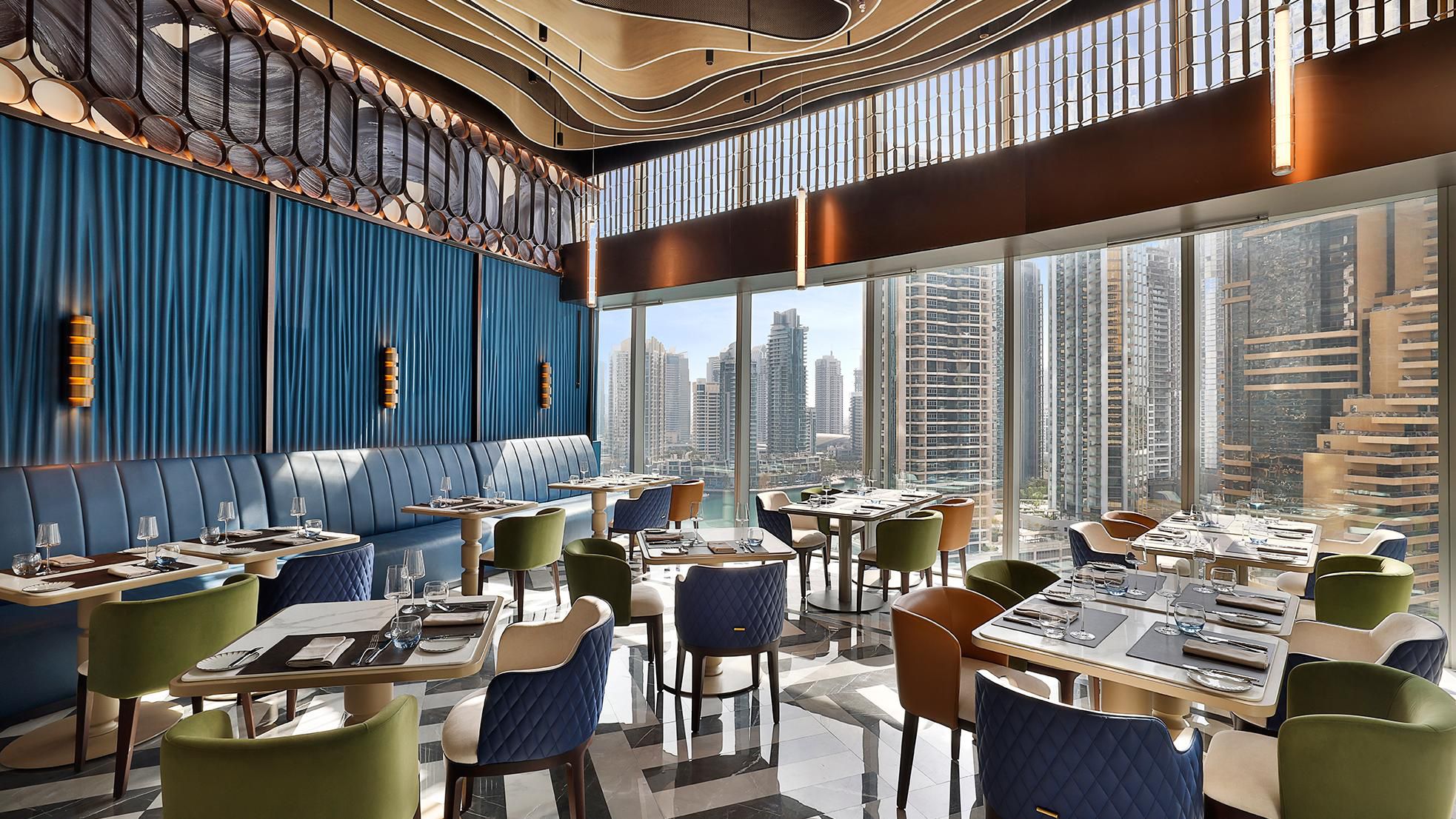 A modern, vibrant dining space featuring a mix of colorful seating geometric flooring, and floor-to-ceiling windows offering expansive views of the Dubai Marina skyline