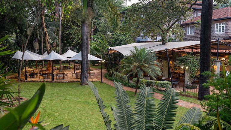 Garden dining area with canopy-covered tables, palm trees, and greenery next to a stone building.