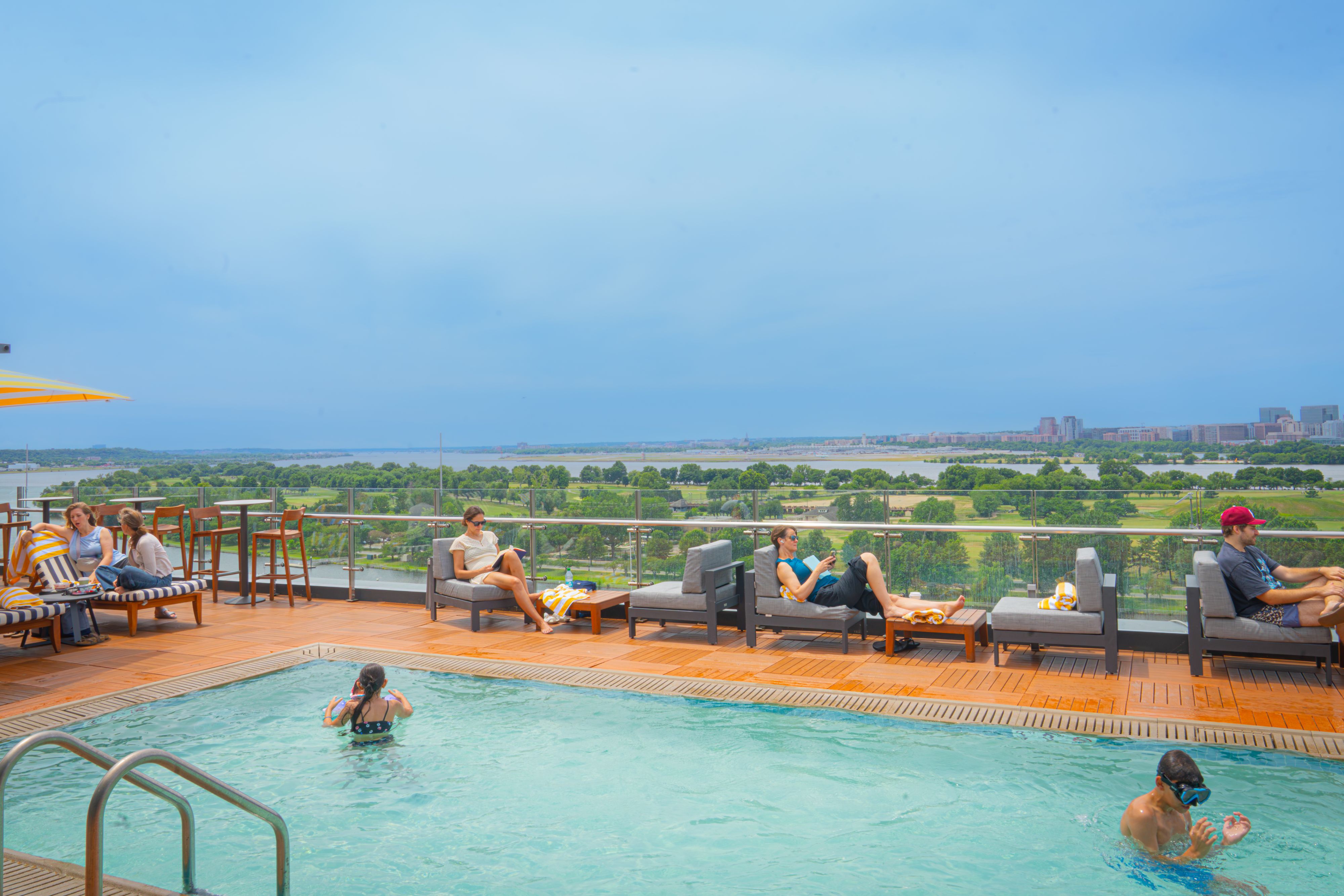 Guests relaxing at a rooftop pool overlooking the Potomac River and Washington, D.C. landmarks on a sunny day.