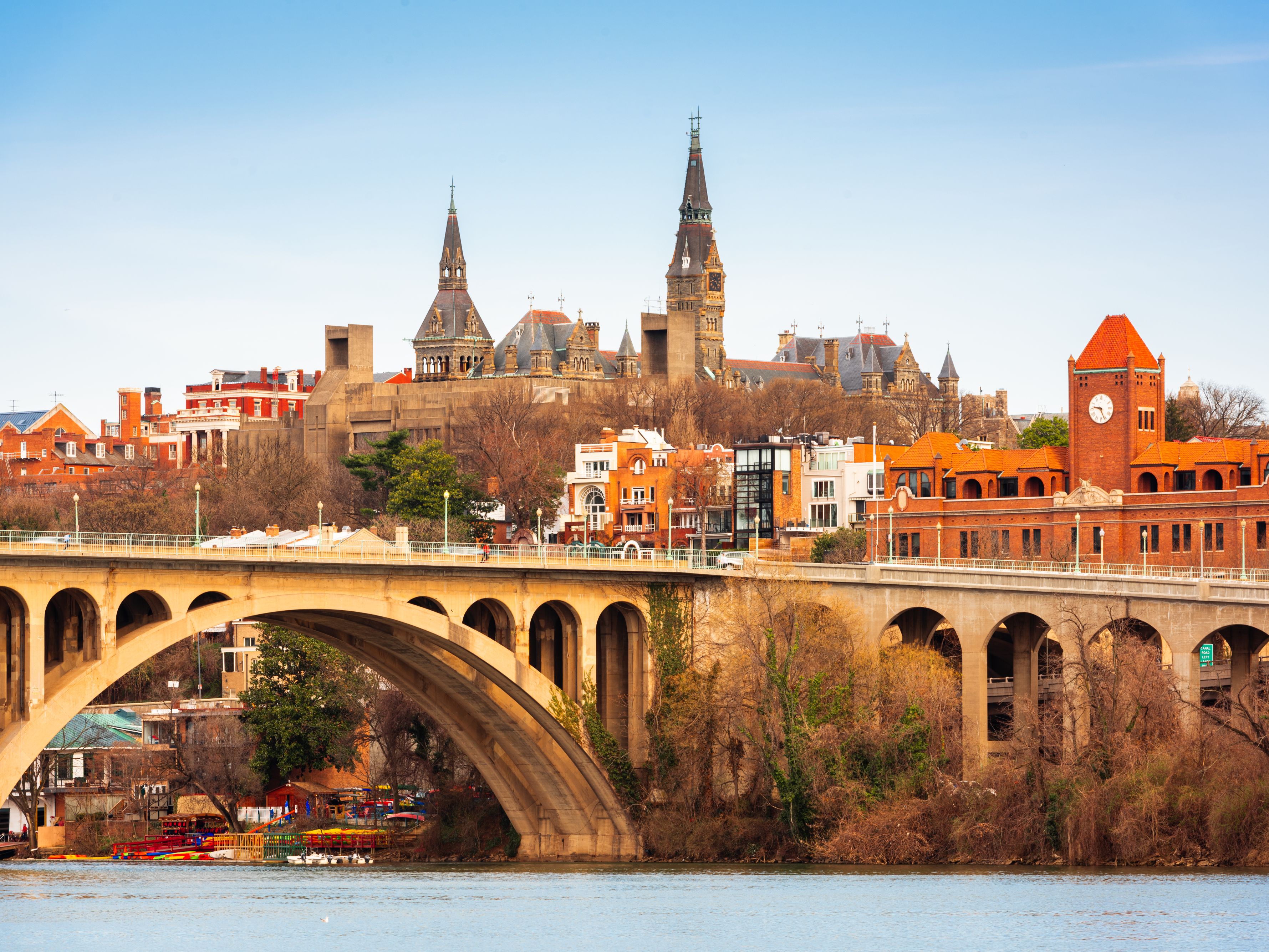 Georgetown waterfront view featuring the arched Key Bridge, historic buildings, and the skyline beyond the Potomac River under a clear blue sky.
