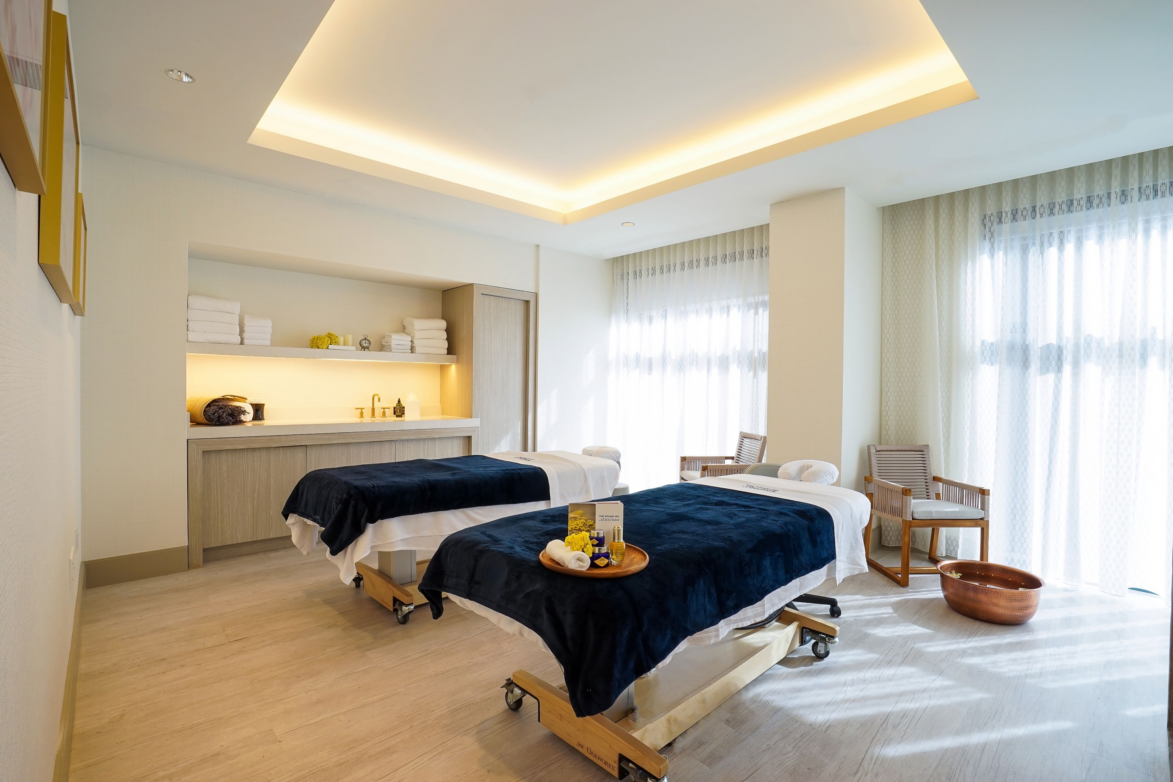Bright spa treatment room with two massage tables, soft linens, natural light, and a calming contemporary design.