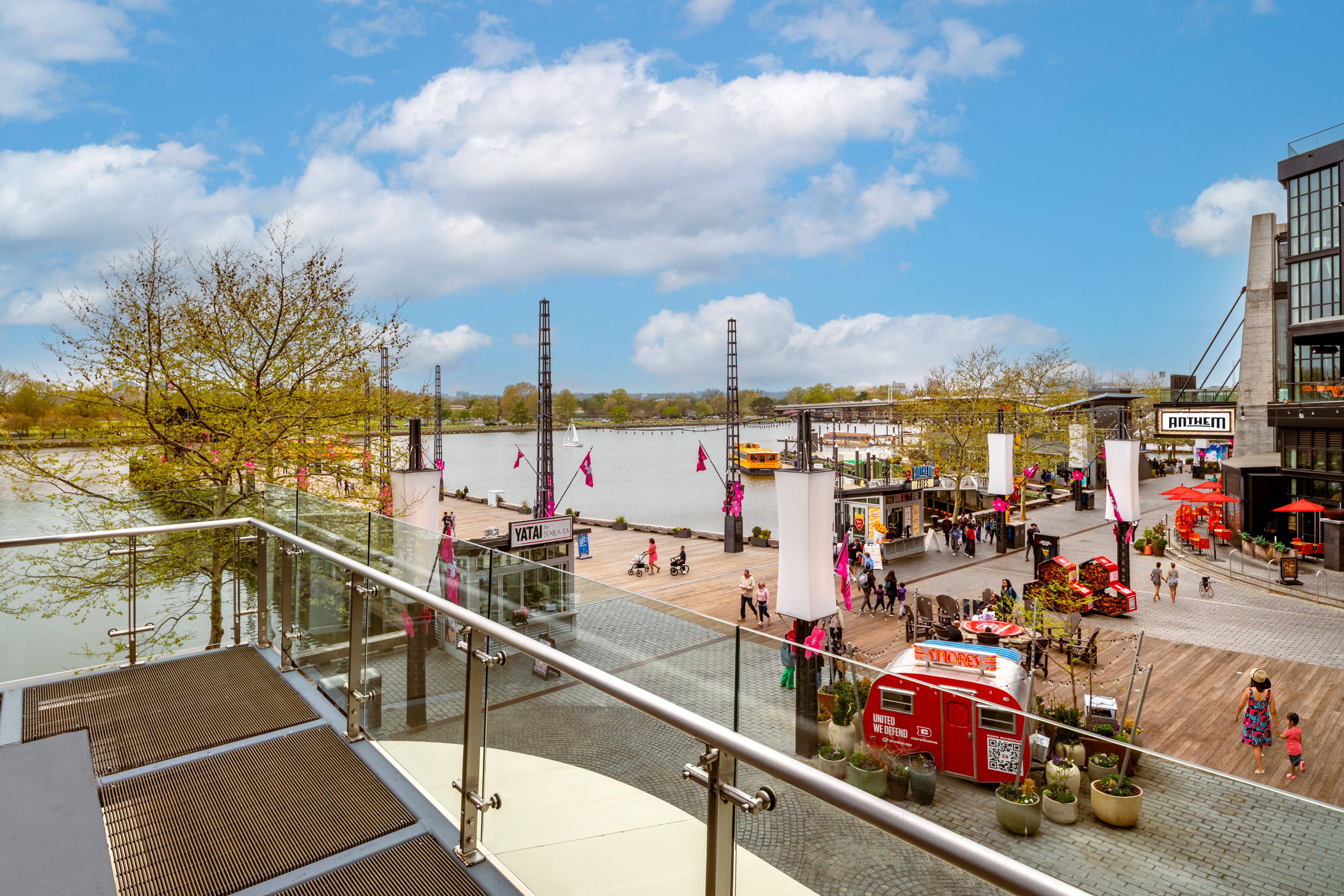 Elevated view of The Wharf waterfront with families strolling along the boardwalk, shops, dining venues, boats, and the Potomac River under a blue sky.
