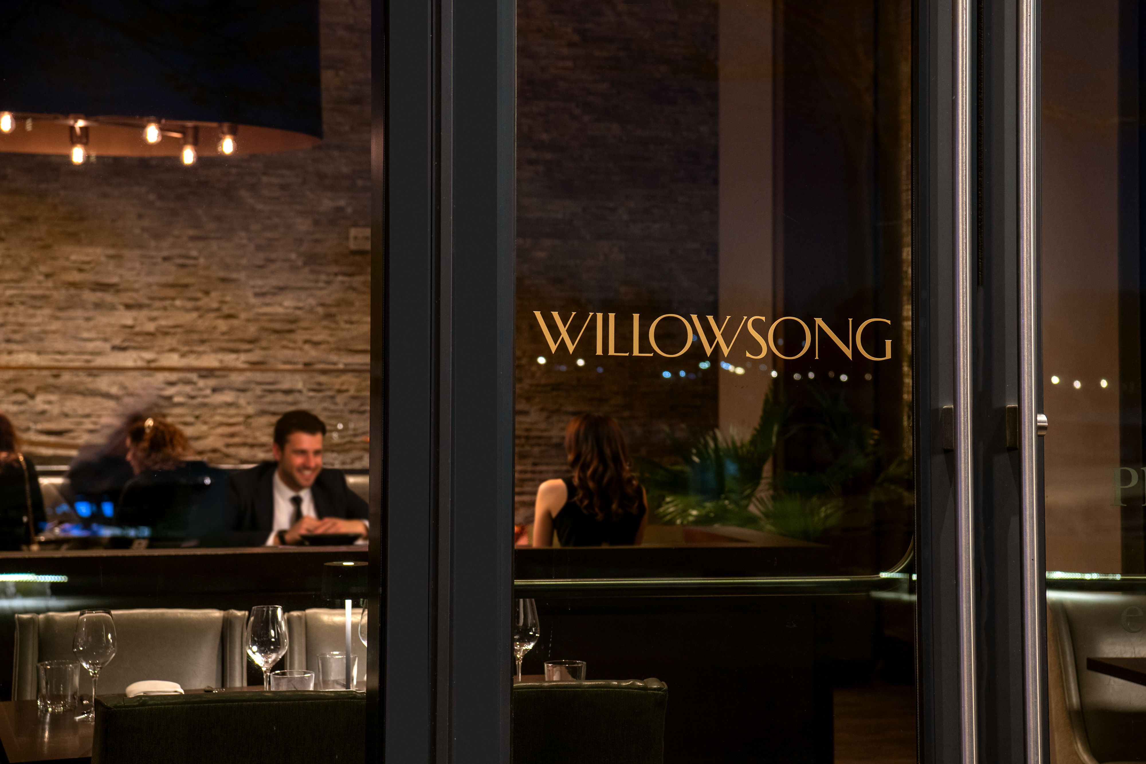Evening view through the glass entrance of Willowsong restaurant, with guests dining in an elegant setting.