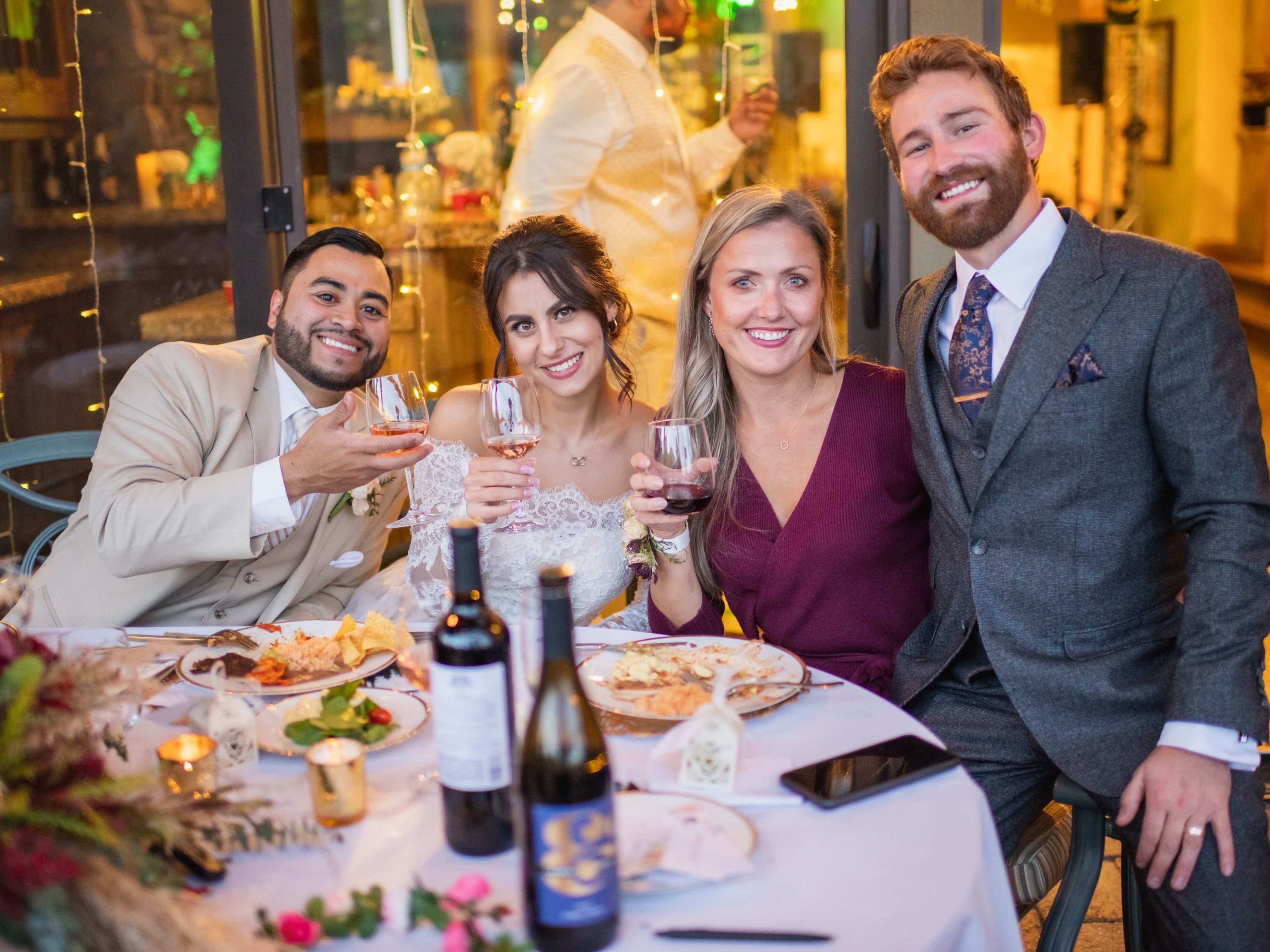 Bride, groom, and friends sitting at an outdoor table with food and wedding decorations.