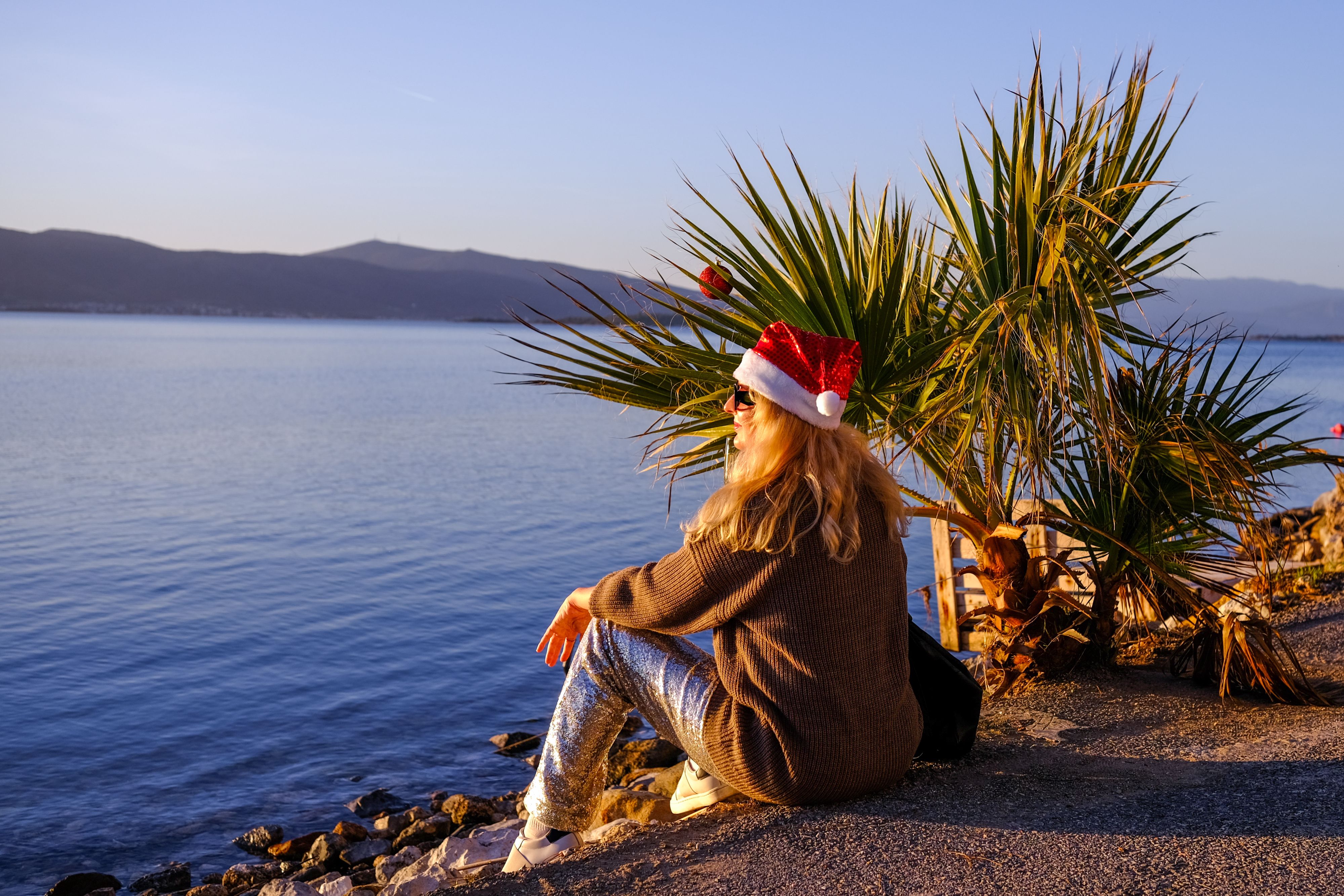 Person wearing a red Santa hat and sunglasses sits on a rocky shoreline beside palm trees, overlooking calm blue water and distant mountains in warm golden-hour light.