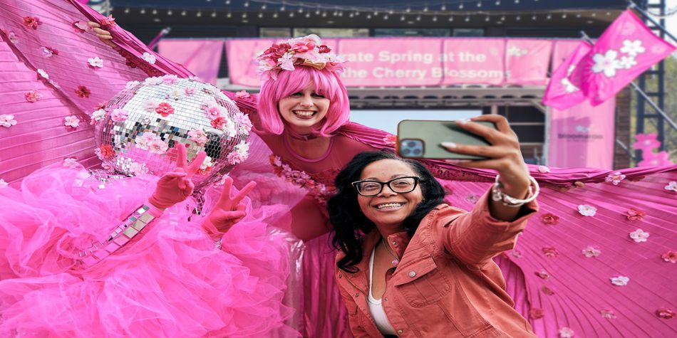 Person taking a selfie with two performers dressed in bright pink costumes decorated with flowers and wings at a cherry blossom-themed event.
