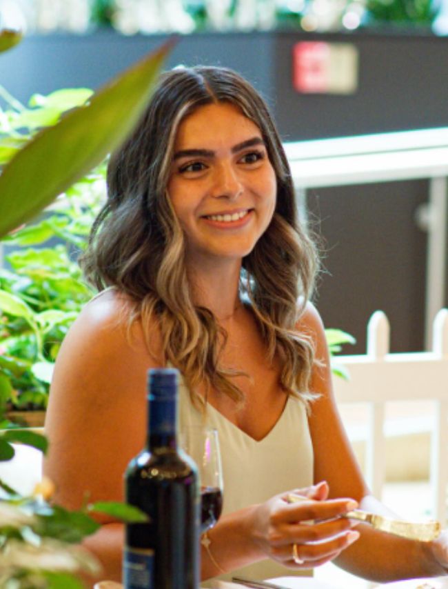 Person seated at a table with a glass of wine and a bottle, surrounded by greenery in a restaurant setting.