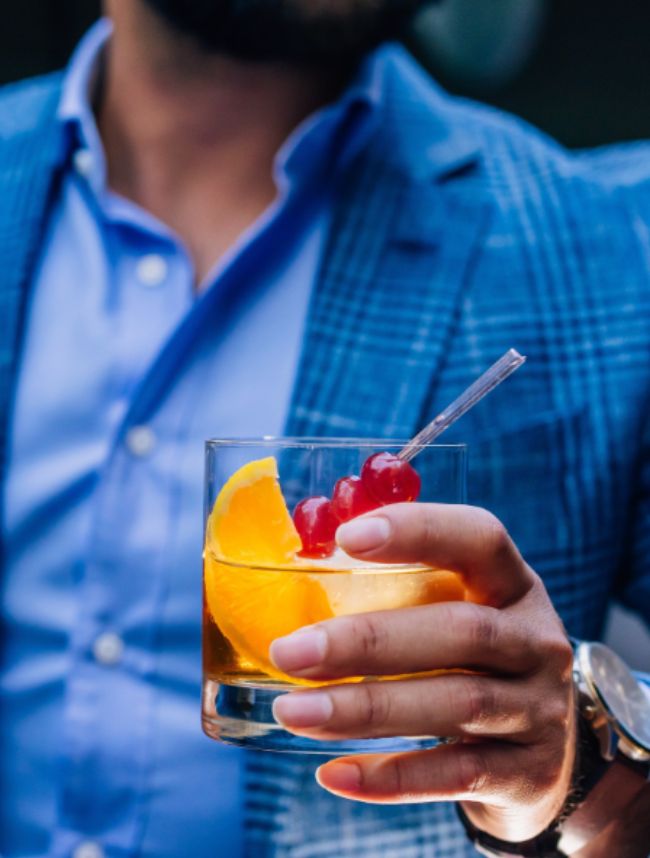 Close-up of a hand holding a cocktail with an orange slice and cherries, with a blue jacket in the background.