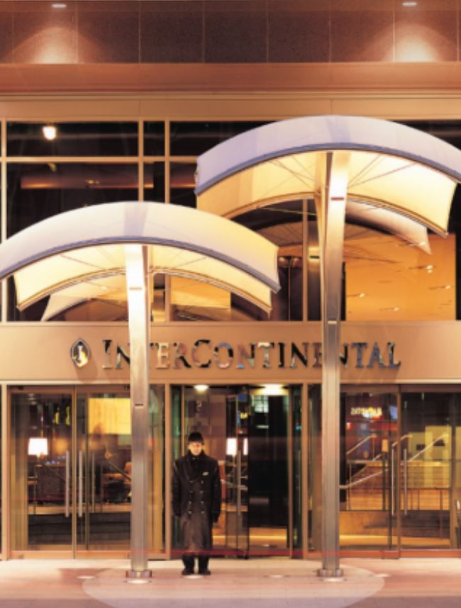 Covered entrance of InterContinental hotel at night with glass doors, signage, and a doorman standing outside.