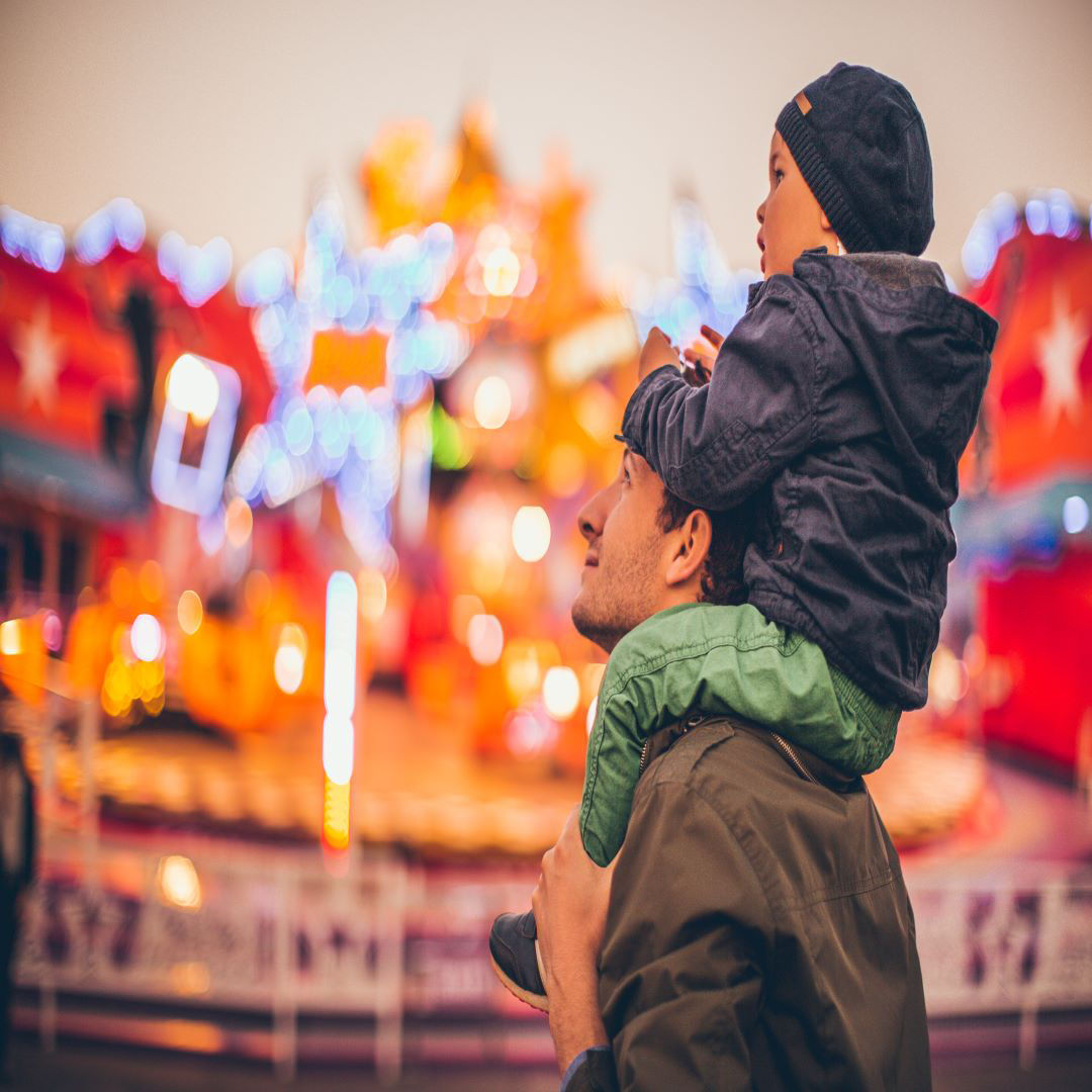 Child on man's shoulders looking at amusement park lights