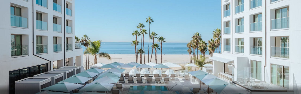 Exterior of the Regent Santa Monica Beach with a pool and beach chairs in the foreground, and the beach and palm trees in the distance.